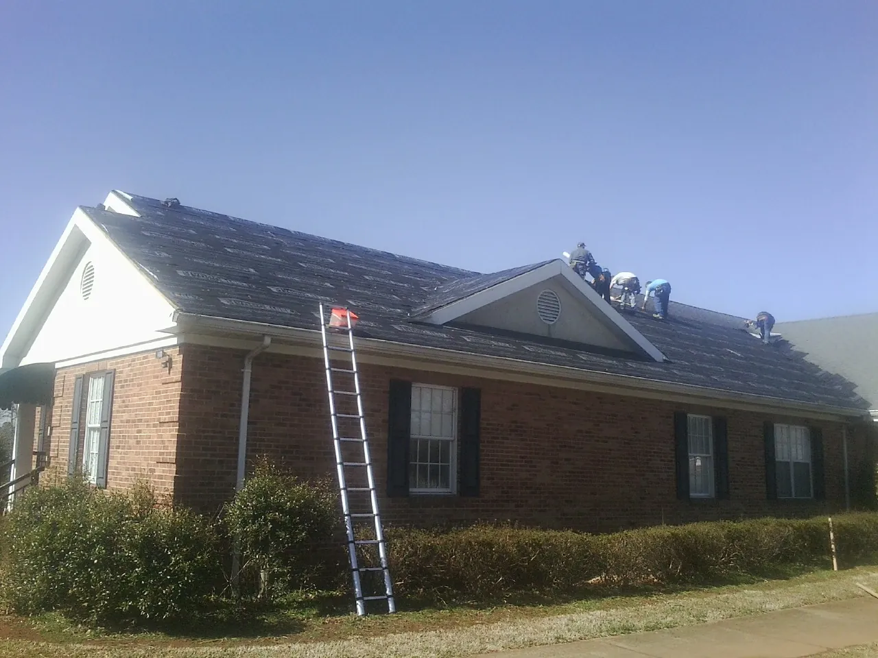 Crew tearing shingles off a brick ranch roof with a ladder set — Emergency Roof Repair work by our crew in Riverdale